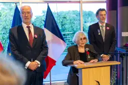 Ceremonia wręczenia odznaczeń, mężczyźni w garniturach, kobieta przy mównicy.