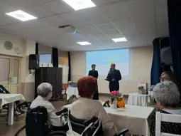 Spotkanie w sali z osobami starszymi, mężczyzna i kobieta na scenie, prezentacja w tle.