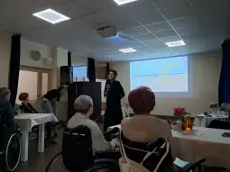 Osoba przemawiająca do grupy ludzi siedzących na wózkach inwalidzkich w sali konferencyjnej.