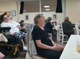 Grupa osób w pomieszczeniu, w tym kobieta siedząca przy stole i osoby w tle.