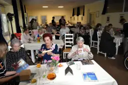 Spotkanie seniorów w sali, przy stołach z napojami i dekoracjami, w atmosferze radości i integracji.