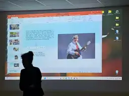 Kobieta z gitarą w tradycyjnym stroju, prezentująca się na tle slajdów.