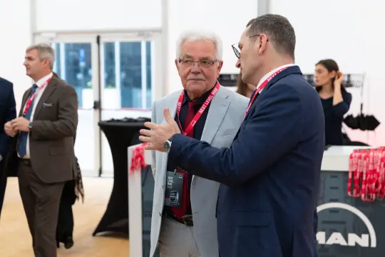 Dwaj mężczyźni rozmawiają w eleganckim otoczeniu, w tle widoczne stoiska i uczestnicy wydarzenia.
