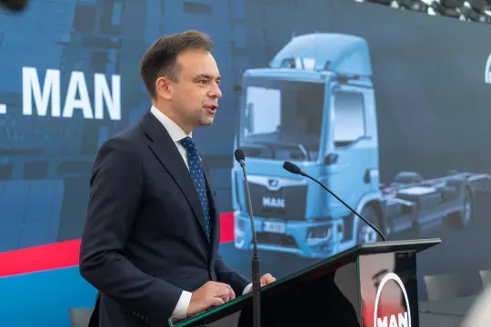 Mężczyzna przemawia przy mikrofonie, w tle widoczny niebieski samochód ciężarowy.