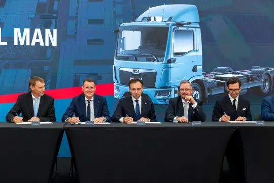 Podpisanie umowy przez pięciu mężczyzn przy stole, w tle grafika ciężarówki.