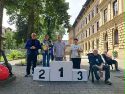 Zwycięzcy na podium, obok nich osoby z nagrodami, w tle budynki i drzewa.
