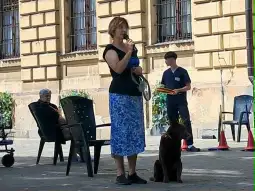 Kobieta z lodami stoi obok psa, w tle osoby siedzące na krzesłach.