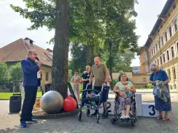 Impreza plenerowa z uczestnikami w wózkach, prowadzący przemawia, w tle budynki i drzewa.