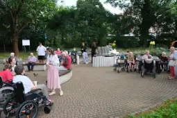 Grupa osób starszych w ogrodzie, część na wózkach inwalidzkich, inne rozmawiają lub odpoczywają.