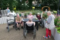 Grupa osób starszych w ogrodzie, siedzących przy stole, otoczonych kwiatami i zielenią.