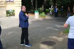 Mężczyzna w niebieskiej marynarce przemawia do grupy ludzi w parku.