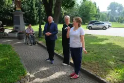 Trzy osoby rozmawiają na świeżym powietrzu, w tle zieleń i samochody.