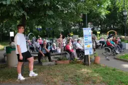 Młody mężczyzna stoi obok grupy seniorów siedzących na ławkach i w wózkach inwalidzkich w parku.