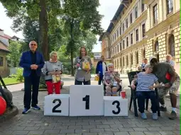 Grupa ludzi na podium, trzymających nagrody, w tle budynki i drzewa.