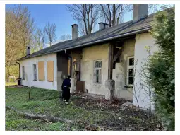 Zniszczony budynek z wybitymi oknami, osoba sprzątająca teren wokół.