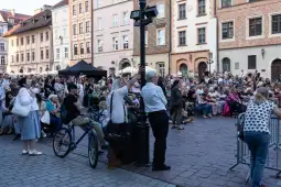 Tłum ludzi zgromadzony na ulicy, niektórzy siedzą, inni stoją, w tle zabytkowe budynki.