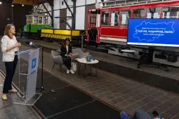 Kobieta przemawia przy stole, obok siedzą dwie osoby, w tle ekran z mapą i tekstem o nowej trasie.
