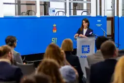 Kobieta przemawia na konferencji, w tle niebieski tramwaj i logo Krakowa.
