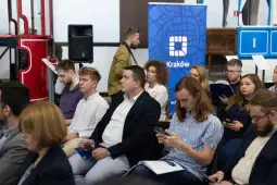 Grupa ludzi siedzących na wydarzeniu, niektórzy korzystają z telefonów. W tle logo Krakowa.