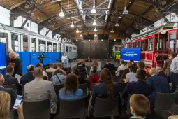 Sala konferencyjna z uczestnikami, tramwaje w tle, ekran z prezentacją.