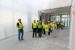 Grupa ludzi w kamizelkach odblaskowych i kaskach na budowie, obserwująca prace remontowe.
