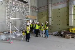 Grupa osób w kamizelkach odblaskowych i kaskach na budowie, przy rusztowaniu.