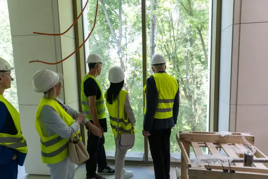 Grupa osób w kaskach i kamizelkach odblaskowych obserwuje widok przez duże okno.