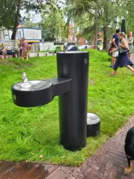 Fontanna do picia w parku, otoczona zieloną trawą i spacerującymi ludźmi.