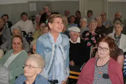 Spotkanie grupy seniorów w sali, kobieta w dżinsowej kurtce uśmiecha się do zgromadzonych.