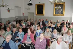 Grupa seniorów uczestniczących w wydarzeniu w sali z obrazami na ścianach.