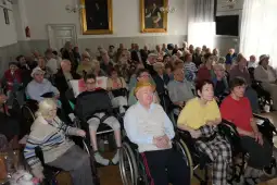 Grupa seniorów w wózkach inwalidzkich uczestniczy w wydarzeniu w sali.