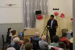 Mężczyzna przemawia do grupy seniorów w sali z dekoracjami kwiatowymi.