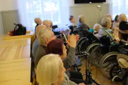 Grupa starszych osób w sali, niektórzy na wózkach inwalidzkich, klaska w dłonie.