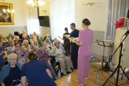 Spotkanie osób starszych z pracownikami opieki w sali, w tle muzyka i aktywności.