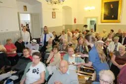 Grupa seniorów w sali, niektórzy jedzą, inni rozmawiają, personel serwuje poczęstunek.