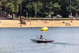 Mężczyzna w łodzi motorowej na wodzie, w tle plaża i ludzie opalający się.