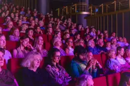 Publiczność w teatrze, skupiona na występie, siedząca na czerwonych fotelach.