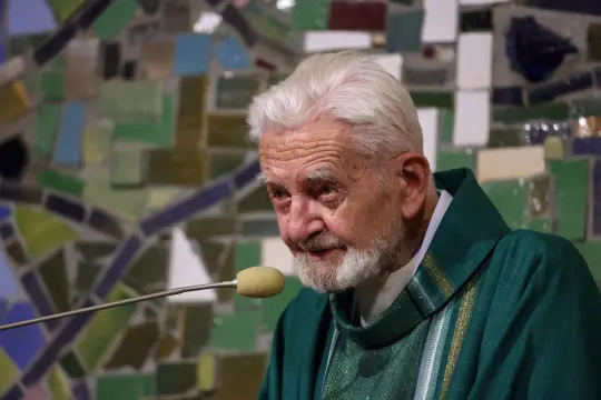 Starszy mężczyzna w zielonym ornacie, przemawiający do mikrofonu. Tło z kolorowych mozaik.