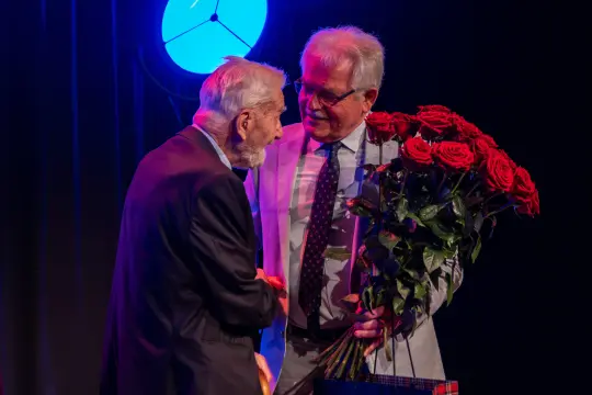 Dwaj mężczyźni wymieniają uściski dłoni, jeden trzyma bukiet czerwonych róż.