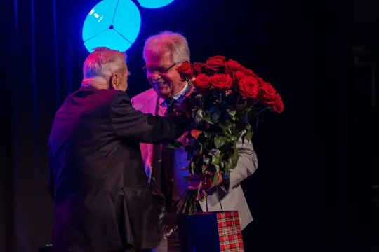 Dwaj mężczyźni wymieniają uściski, jeden trzyma bukiet czerwonych róż.