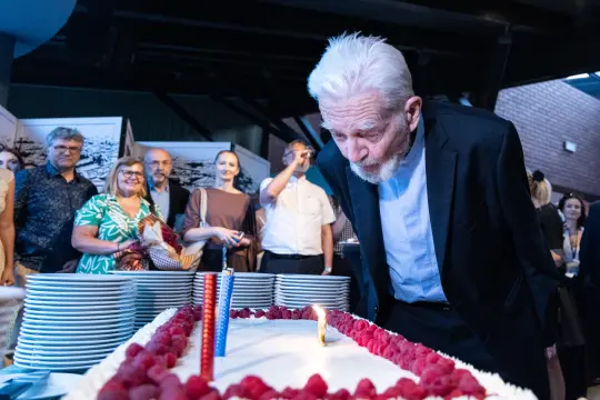 Mężczyzna dmucha na świeczki na torcie urodzinowym otoczony gośćmi.