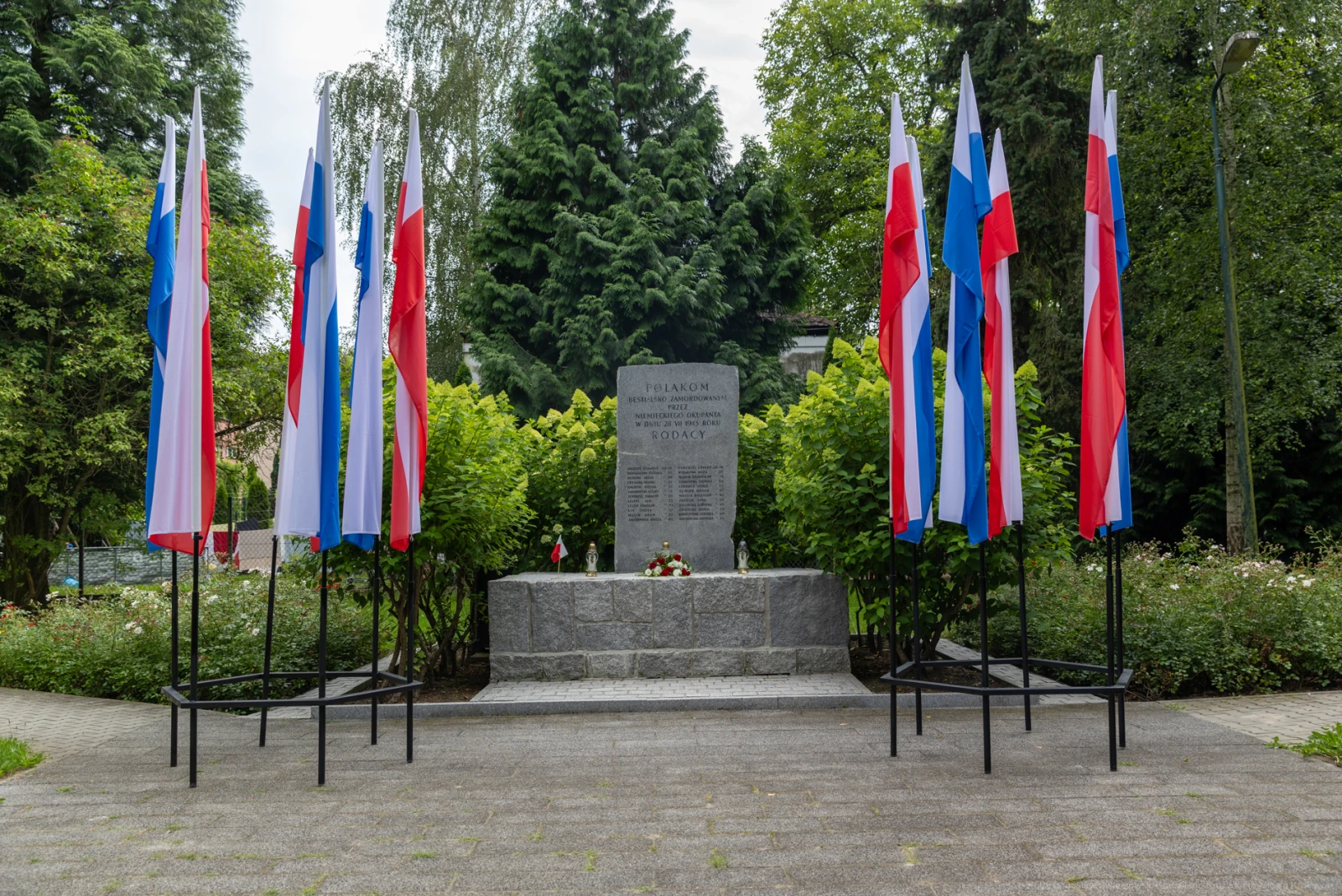 Pomnik z flagami w kolorach narodowych, otoczony zielenią i kwiatami.  Autor: Bogusław Świerzowski