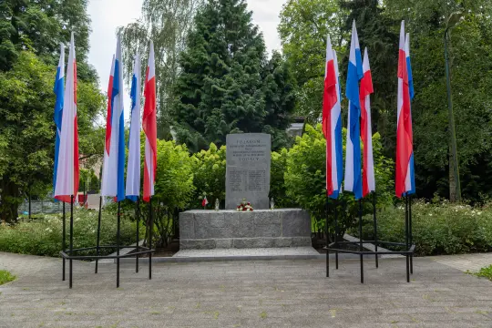 Pomnik z flagami w kolorach narodowych, otoczony zielenią i kwiatami.