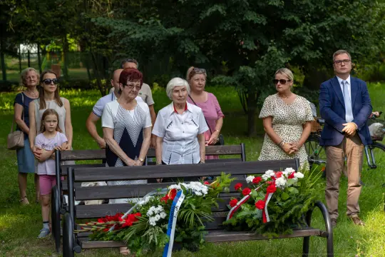Grupa ludzi stojąca przy ławce z kwiatami, uczestnicząca w uroczystości upamiętniającej.