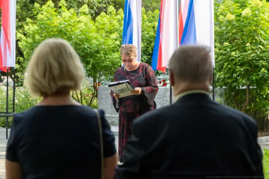 Kobieta czyta tekst w otoczeniu flag i zieleni, dwie osoby siedzą w tle.