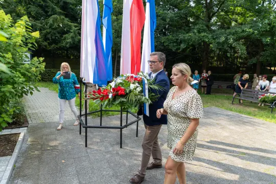 Mężczyzna i kobieta niosą kwiaty w otoczeniu flag, w tle widoczni inni uczestnicy wydarzenia.