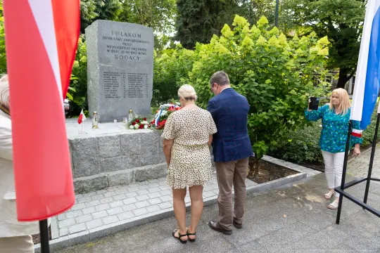 Uroczystość przy pomniku z kwiatami i flagami, dwie osoby składają hołd.