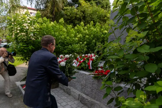 Mężczyzna składa kwiaty przy pomniku, w tle zielone krzewy.