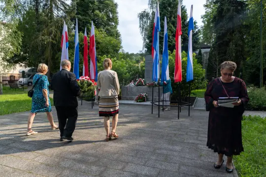 Uczestnicy ceremonii składają kwiaty przy pomniku, w tle flagi narodowe.