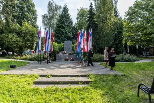 Uroczystość przy pomniku z flagami Polski i uczestnikami w tle.